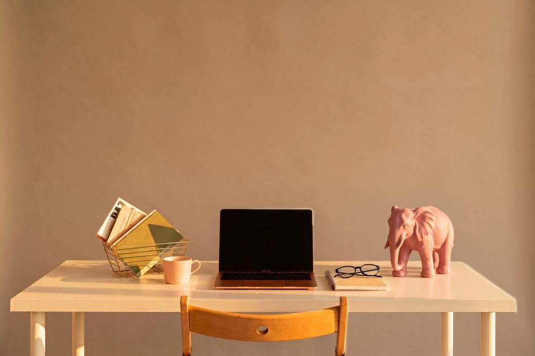The same desk in warm golden-hour light with soft shadows, the potted plant now a pink elephant figurine — after editing with AI Image Editor.