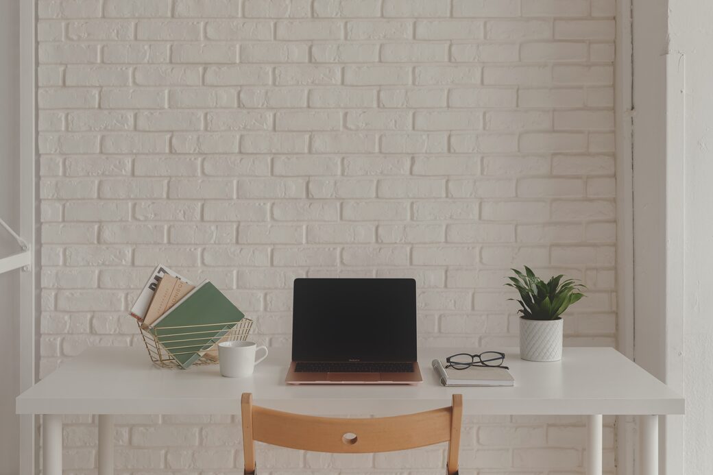 A home-office desk — laptop, coffee mug, notebooks and glasses — against a white brick wall in flat, even light, before editing.